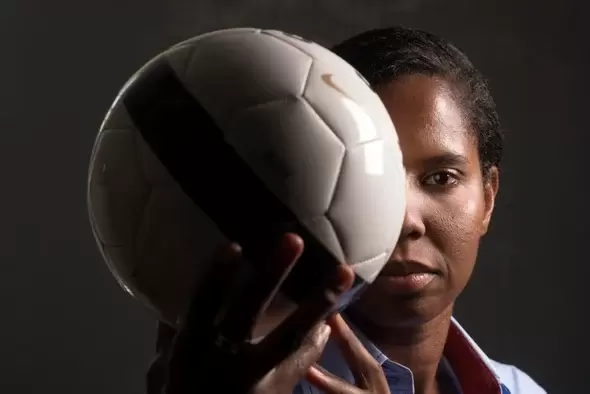 a black female soccer player, briana scurry, holds a soccer ball in front of her face under dramatic lighting. she is a two-time olympic gold medalist and world cup champion. she suffered a traumatic brain injury in 2010 that ended her career