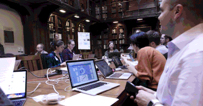 a group of people sit around a table with laptops. behind them are shelves of books. the image flickers with different colors, an artistic design of digital decay