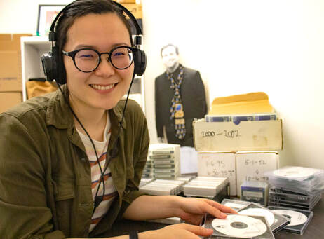 An Asian American woman with black rimmed glasses and headphone smiles next to a desk filled with tapes and CDs