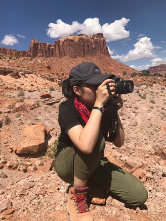 A woman in a baseball hat is kneeling and holding a camera up to her face to take a photo out in a desert landscape